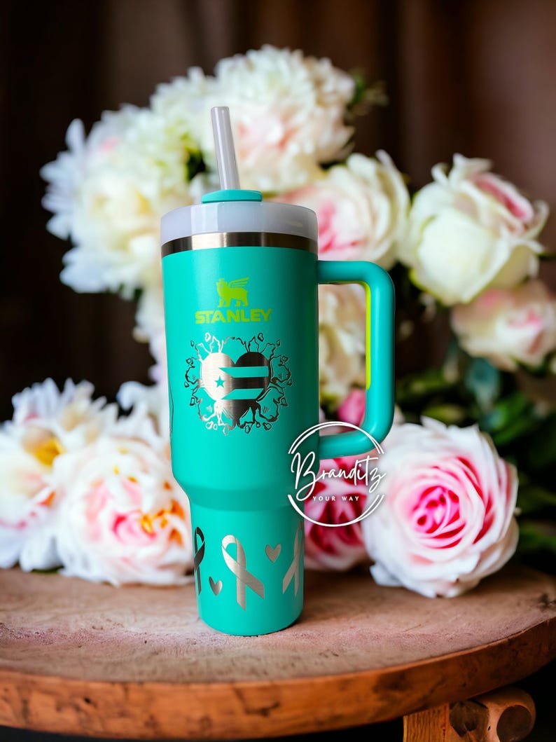 Op de afbeelding: Een turquoise Stanley-beker met een handvat en een rietje. De beker heeft een zilveren ontwerp van een hart, linten en het Stanley-logo. De handgreep en het deksel zijn licht turquoise van kleur. De achtergrond bevat witte en roze bloemen.