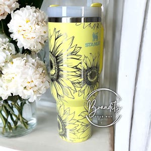 May include: A yellow Stanley tumbler with a sunflower design. The tumbler has a silver rim and a clear straw. The Stanley logo is visible on the side. The tumbler is decorated with black line art sunflowers.
