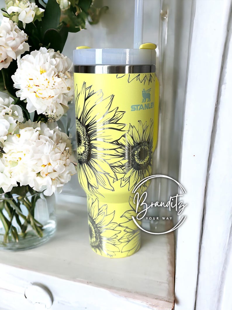 May include: A yellow Stanley tumbler with a sunflower design. The tumbler has a silver rim and a clear straw. The Stanley logo is visible on the side. The tumbler is decorated with black line art sunflowers.