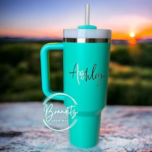 Op de afbeelding: Een turquoise beker met een handvat, een wit deksel en een doorzichtig rietje. De naam "Ashley" staat in sierlijke letters op de voorkant. De beker heeft een gouden band aan de bovenkant en het logo "Branditz Your Way" aan de onderkant.