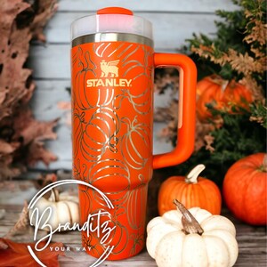 Op de afbeelding: Een oranje Stanley-beker met een pompoenontwerp. De beker heeft een handvat en een doorzichtig deksel. Het Stanley-logo is zichtbaar. De beker is versierd met een zilveren pompoenpatroon. Pompoenen en herfstbladeren omringen de beker.