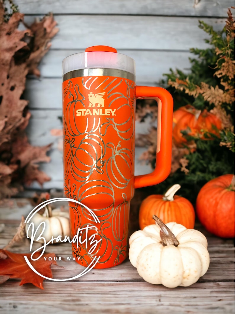 Op de afbeelding: Een oranje Stanley-beker met een pompoenontwerp. De beker heeft een handvat en een doorzichtig deksel. Het Stanley-logo is zichtbaar. De beker is versierd met een zilveren pompoenpatroon. Pompoenen en herfstbladeren omringen de beker.