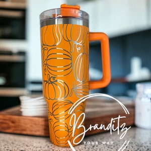 Könnte beinhalten: Ein orangefarbener Reisebecher mit einem silbernen Kürbismuster und einem orangefarbenen Griff. Der Becher hat einen durchsichtigen Deckel und den Text "Branditz YOUR WAY" in weißer Schrift. Der Becher steht auf einem Holztablett.