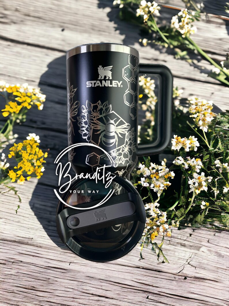 Op de afbeelding: Zwarte Stanley reismok met een bijen- en bloemenontwerp. De mok heeft een handvat en een deksel. De tekst "STANLEY" is zichtbaar op de mok. De mok is versierd met een bijen- en honingraatpatroon.