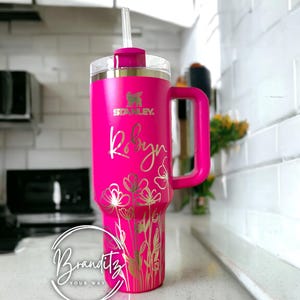 Op de afbeelding: Een felroze Stanley-beker met een handvat en een doorzichtig rietje. De beker is gepersonaliseerd met de naam "Robyn" in gouden letters, samen met een bloemenontwerp. De beker heeft het Stanley-logo en het Branditz-logo.