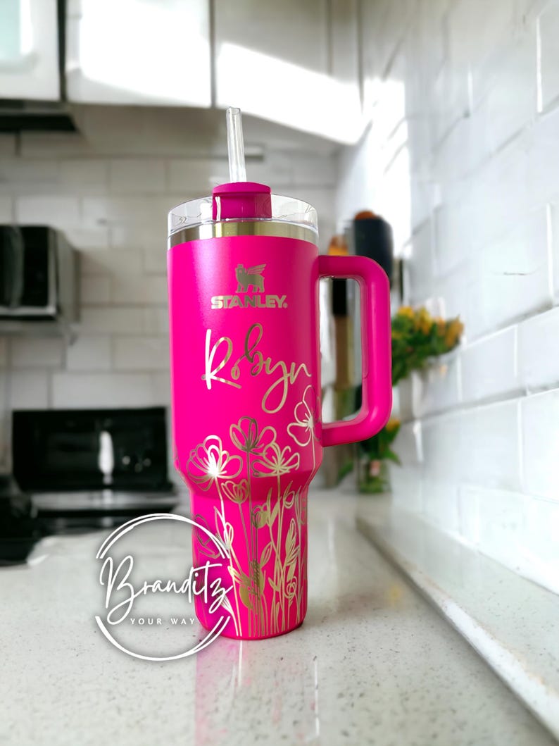 Op de afbeelding: Een felroze Stanley-beker met een handvat en een doorzichtig rietje. De beker is gepersonaliseerd met de naam "Robyn" in gouden letters, samen met een bloemenontwerp. De beker heeft het Stanley-logo en het Branditz-logo.