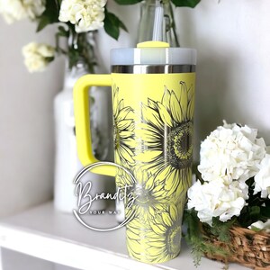 May include: A yellow tumbler with a sunflower design. The tumbler has a handle and a lid. The text "Branditz YOUR WAY" is in a circle on the front of the tumbler. The tumbler is sitting on a white shelf with flowers in the background.