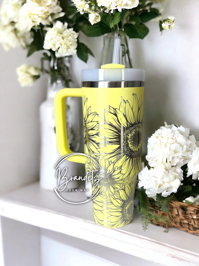 May include: A yellow tumbler with a sunflower design. The tumbler has a handle and a lid. The text "Branditz YOUR WAY" is in a circle on the front of the tumbler. The tumbler is sitting on a white shelf with flowers in the background.