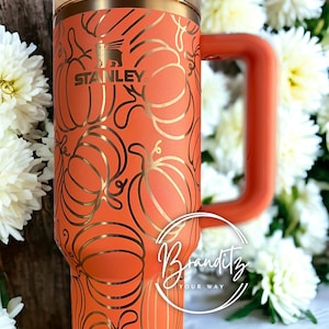 May include: Orange Stanley tumbler with a pumpkin design in gold. The tumbler has a handle and a lid. The Stanley logo is visible. The tumbler is surrounded by white flowers.