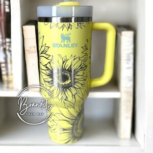 May include: A yellow Stanley tumbler with a sunflower design and a matching yellow handle. The tumbler has a gray lid and a clear straw. The Stanley logo is visible on the front. The tumbler is displayed on a white shelf with books.