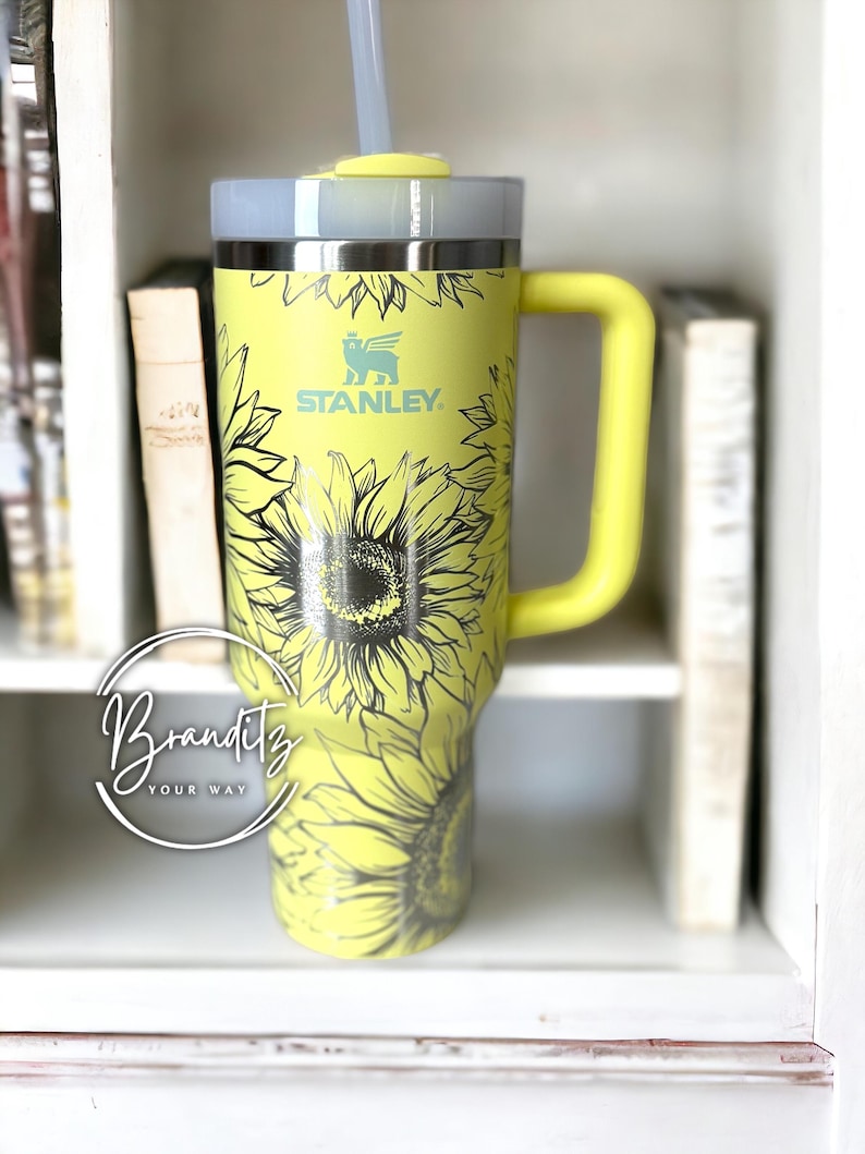 May include: A yellow Stanley tumbler with a sunflower design and a matching yellow handle. The tumbler has a gray lid and a clear straw. The Stanley logo is visible on the front. The tumbler is displayed on a white shelf with books.
