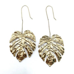 May include: A pair of gold-tone earrings featuring a large, detailed monstera leaf design. The leaves are textured and have a cutout pattern. The earrings hang from a thin chain and have a hook closure.