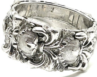 May include: A silver ring with a detailed, ornate design featuring two stylized hearts and intricate floral patterns.