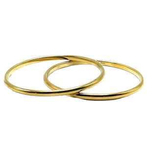 Peut inclure: Deux joncs fins et circulaires en or avec une finition lisse et polie. Les bracelets sont entrelacés, créant une forme qui se chevauche. Le métal présente un ton or jaune brillant et réfléchissant, avec une épaisseur uniforme.