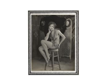 May include: A black and white vintage photograph in a decorative silver frame depicting a person with short hair sitting on a wooden stool. The person wears a halter-style top and shorts, posing with one hand near their face, set against a backdrop.