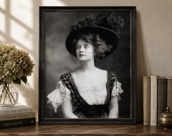 May include: A black and white portrait print in a dark frame featuring a person wearing a wide-brimmed hat with dark feathers and a light-colored blouse with dark, textured straps. The person has dark hair and is looking toward the upper left.