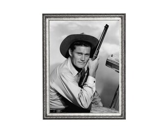 May include: Black and white framed portrait of a man in a cowboy hat holding a rifle. The man is wearing a light-colored button-down shirt and looking towards the camera. The frame is silver.
