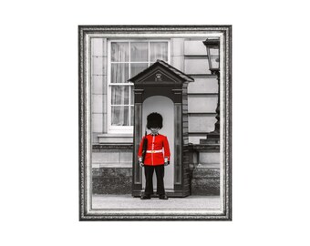 May include: Framed black and white photograph of a British Royal Guard in a red uniform and black bearskin hat, standing in a sentry box. The background is a building with a window and a street lamp.