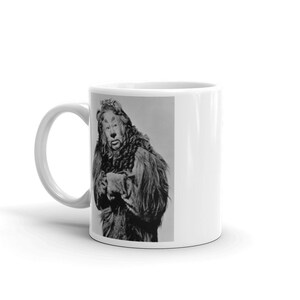 May include: A white ceramic mug with a black and white image of the Cowardly Lion from the Wizard of Oz. The lion is wearing a fur coat and has a human face.