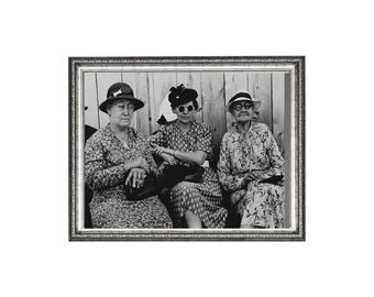 May include: Black and white framed photograph of three women seated together. They are wearing hats and patterned dresses. The photograph is in a silver frame and is a vintage portrait.