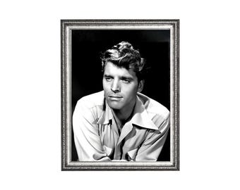 May include: Black and white framed portrait of a man with styled hair, wearing a light-colored button-down shirt. The portrait is in a silver-colored frame, set against a dark background. The man is looking off to the side.