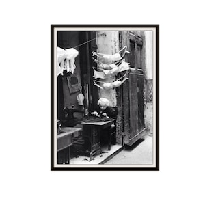 以下が含まれることがあります： 小さな部屋のテーブルで縫い物をしている女性の白黒写真。彼女の頭上には、白い衣服が何枚か紐にかけられています。