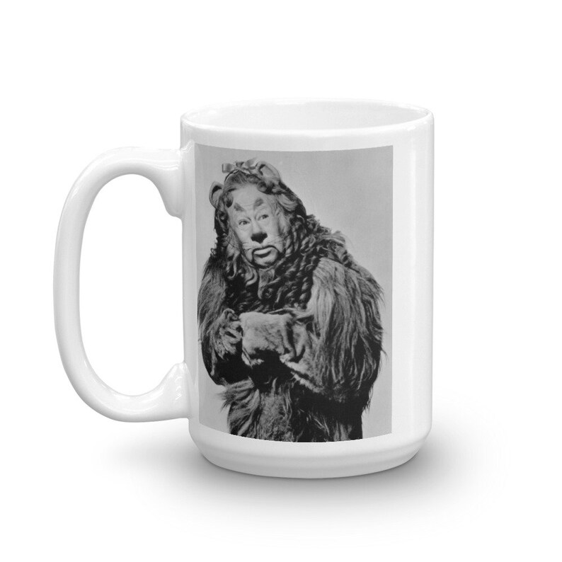 May include: A white ceramic mug with a black and white image of the Cowardly Lion from the Wizard of Oz. The lion is wearing a fur coat and has a serious expression on its face.
