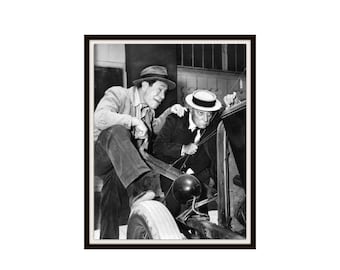 May include: A black and white photograph featuring two individuals in vintage attire examining the engine of an antique automobile. One individual wears a fedora and a light-colored jacket, while the other wears a straw boater hat and a dark suit.