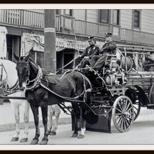 May include: A black and white vintage fire engine with two horses pulling it. The fire engine has a large water tank and a ladder on the back. The horses are wearing harnesses and have bridles on their heads. The fire engine is on a paved road.