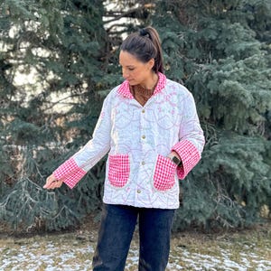 May include: White quilted jacket with pink gingham trim on the cuffs, collar, and pockets. The jacket features floral embroidery and a button-up front. The person is wearing black jeans.