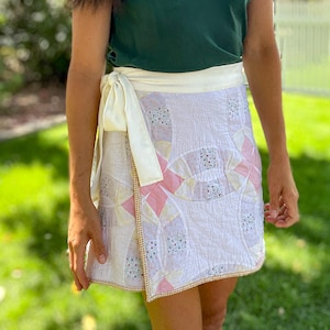 Peut inclure: Jupe portefeuille en patchwork avec des motifs roses, jaunes et floraux sur fond blanc. La jupe a une fermeture à nouer crème et une bordure dorée. La personne porte un caraco vert foncé. La jupe est faite à partir d'une courtepointe.