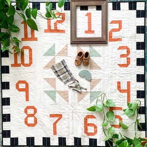 May include: A white quilt with black and white squares around the border. The quilt has a patchwork design with numbers 1 through 12 in orange. There are also two small green and brown triangles in the center of the quilt.