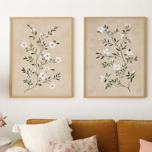 May include: Two framed botanical prints with white flowers and green leaves on a beige background hang above a mustard-colored sofa. The sofa is adorned with pillows in various textures and patterns, including floral and solid pink. A side table with a lamp and vase of flowers completes the scene.