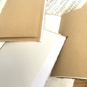 May include: A stack of brown paper notebooks with white edges. The notebooks are bound with white stitching and have a rustic, handmade look.
