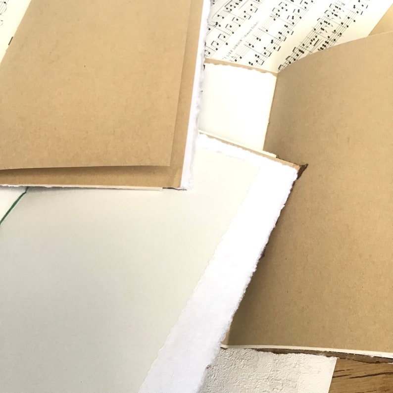 May include: A stack of brown paper notebooks with white edges. The notebooks are bound with white stitching and have a rustic, handmade look.