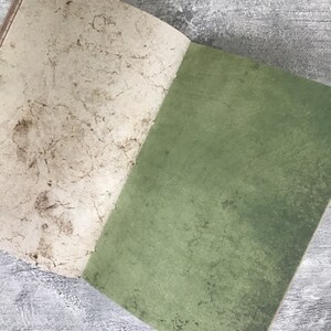 Puede incluir: Un cuaderno abierto con una cubierta texturizada de color blanquecino y una cubierta verde s&oacute;lida. El cuaderno est&aacute; acostado sobre una superficie gris.
