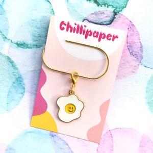 Puede incluir: Marcapáginas dorado con un amuleto de huevo frito de esmalte blanco con una carita sonriente amarilla. El marcapáginas está en una tarjeta rosa y blanca con el texto "Chillipaper" en rosa.