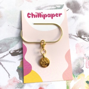 May include: Gold-toned metal bookmark with a paw print charm and a hook to attach to a page. The bookmark is on a pink and yellow background with the text "Chillipaper" printed on the card.