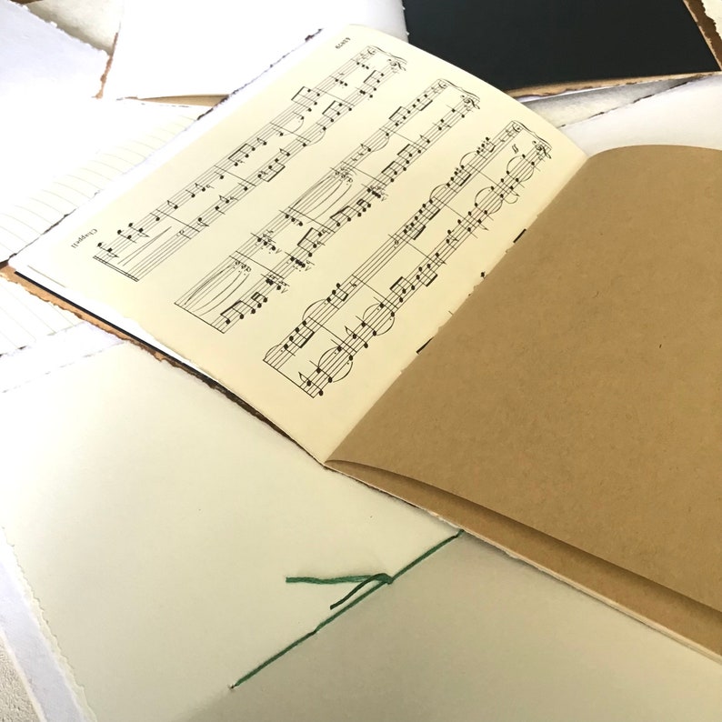 May include: A brown leather-bound journal with a sheet of music paper inside. The journal is tied closed with a green string.