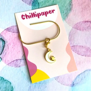 May include: Gold bookmark with a white crescent moon and a gold star charm. The bookmark is on a pink and yellow card with the text "Chillipaper" printed on it.