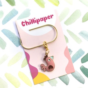 May include: A gold stitch marker with a pink chicken charm. The charm has a white eye and a blue beak. The card reads "Chillipaper".