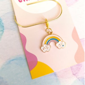 Könnte beinhalten: Ein goldener Anhänger mit einem Regenbogen und Wolken. Der Regenbogen hat gelbe, orange, lila und blaue Streifen. Die Wolken sind weiß mit einem hellblauen Umriss und haben ein Smiley-Gesicht.