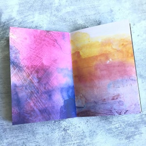 May include: An open notebook with two pages painted in watercolour. The left page is a blend of pink, purple, and blue, while the right page features a yellow, orange, and red sunset-like design.