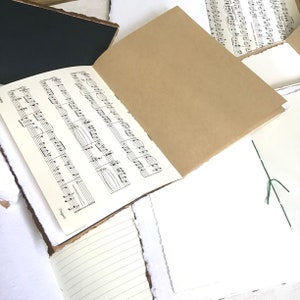 May include: A brown paper notebook with a sheet of music paper inside. The notebook has a rough, textured cover and is open to reveal the music paper. The music paper has a black ink score with the word "Cappelli" printed at the bottom.