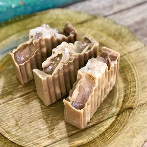 May include: Three bars of handmade soap with a brown and beige color. The soap has a rough texture and is topped with white sea salt.