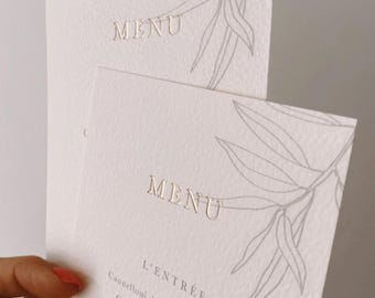 Puede incluir: Una tarjeta de menú blanca con letras doradas y un diseño floral gris. El menú presenta el texto "MENU", "L'ENTRÉE", "Cannelloni de Seiches & Gambas", "Cappuccino de langoustine", "LE PLAT", "Pièce de cochon taillée dans la", "Label Rouge du Lim" y "Polenta crémeux".