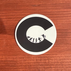 May include: A black and white sticker with a stylized letter "C" that has a circle inside of it. The circle contains a silhouette of the evolution of humans, starting with a monkey and ending with a person holding a surfboard.