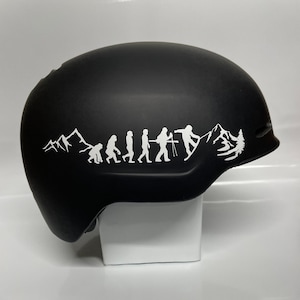 Puede incluir: Casco de snowboard negro con un gráfico blanco que representa la evolución de un snowboarder. El gráfico muestra una progresión de simio a snowboarder, con siluetas de montañas y árboles. Casco diseñado para deportes de invierno.