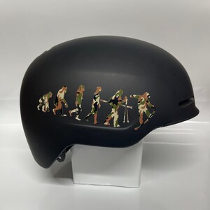 May include: Black ski helmet with a camouflage evolution of man sticker. The sticker shows a series of figures progressing from a primitive ape to a skier.