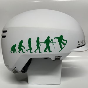 May include: White ski helmet with a green evolution of man sticker on the side. The sticker shows a series of stick figures progressing from a monkey to a skier. The helmet has the word "SMITH" printed on the side.